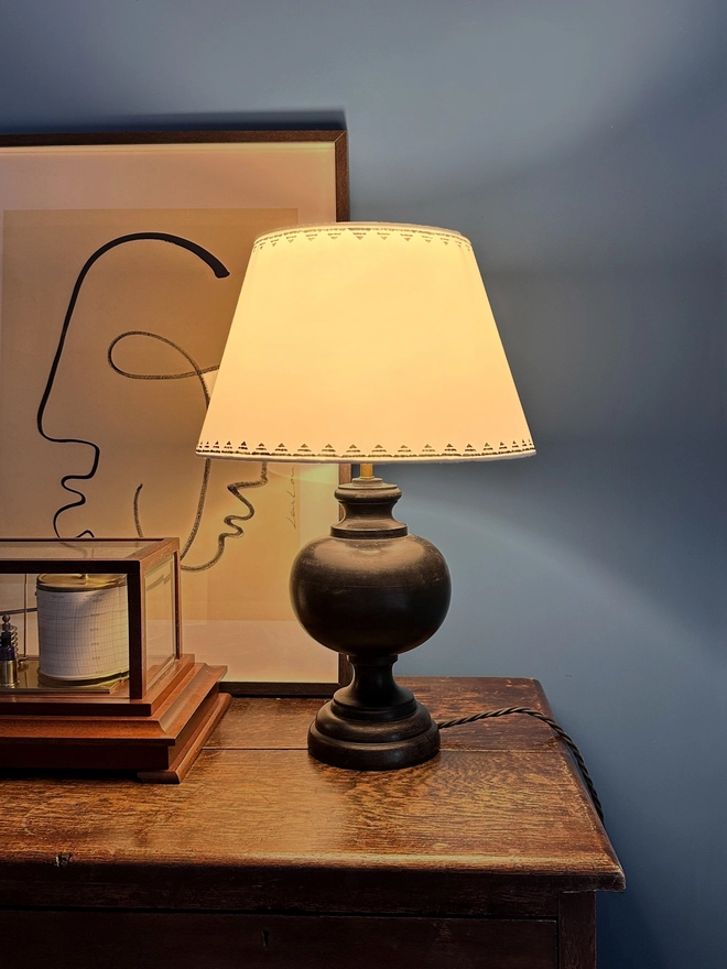 Romantic evening setting with a glowing cream empire lampshade featuring a lined triangle border in dark ink blue. The geometric lampshade casts a warm light, styled on a dark wooden lamp base against a deep blue wall.