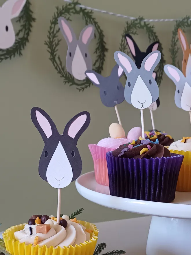 Dark brown and white easter bunny cake topper in iced cupcake. Cake stand in background holds selection of cupcakes with other bunny cupcake toppers