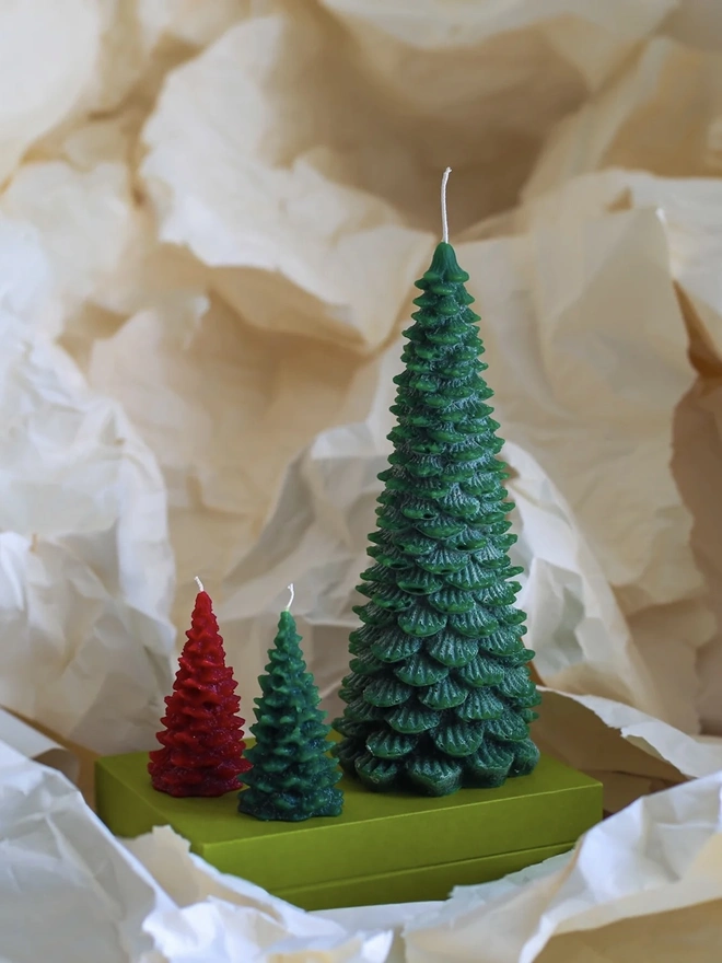 Large Christmas Tree Candle. a large dark green christmas tree shaped candle stands on a green box, nestled among crumpled cream tissue paper. a smaller red and a smaller lighter green tree candle sit in front of it for scale, both with visible wicks. the textured, layered pine branch design and slightly variegated wax give all three candles a realistic, hand-poured finish.