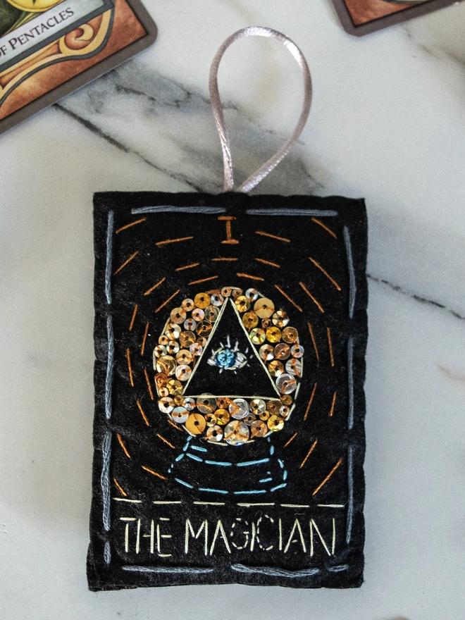 A handcrafted felt ornament with sequins and embroidery depicting "The Magician" tarot card. Colourful embroidery with an eye inside a crystal ball. 