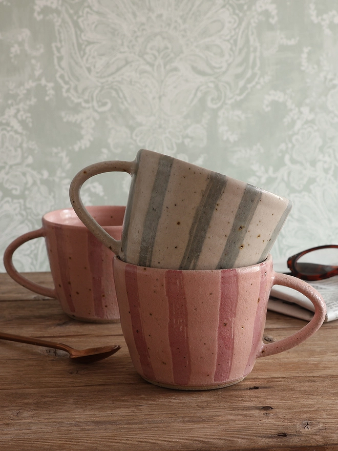 Handmade Ceramic Stoneware Mug in Blue and White Stripe stacked on top of our Handmade Ceramic Stoneware Mug in Pink Stripe, sat on a wooden surface with another pink stripe mug, a newspaper and glasses, and a brass spoon. 