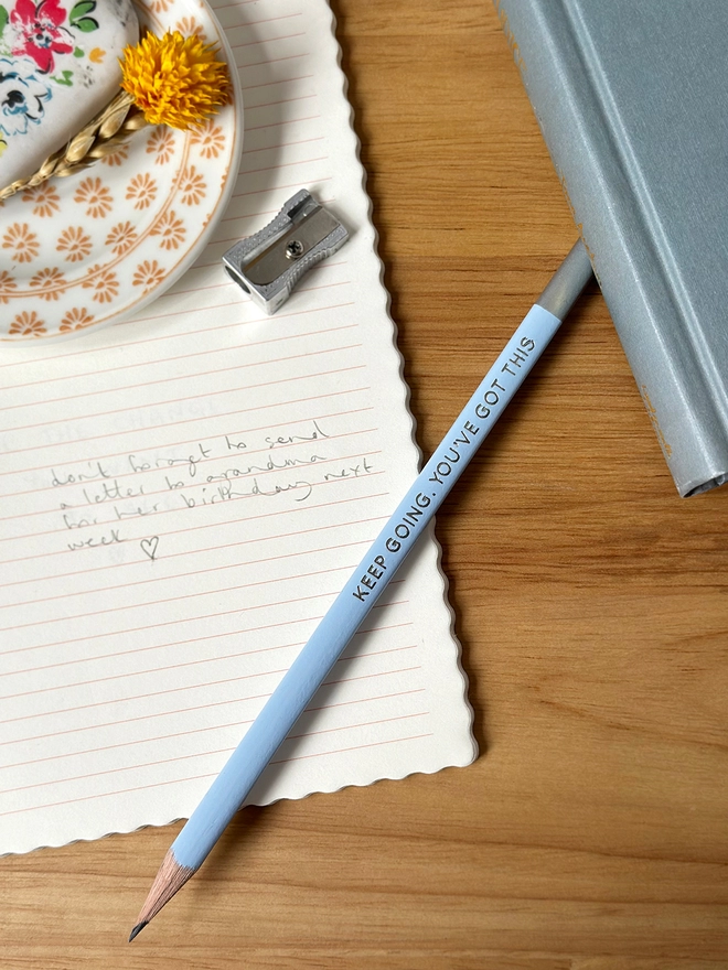 A blue pencil with the words Keep Going, You've Got This in silver writing on the side rests on an open lined notebook both on a wooden desk.