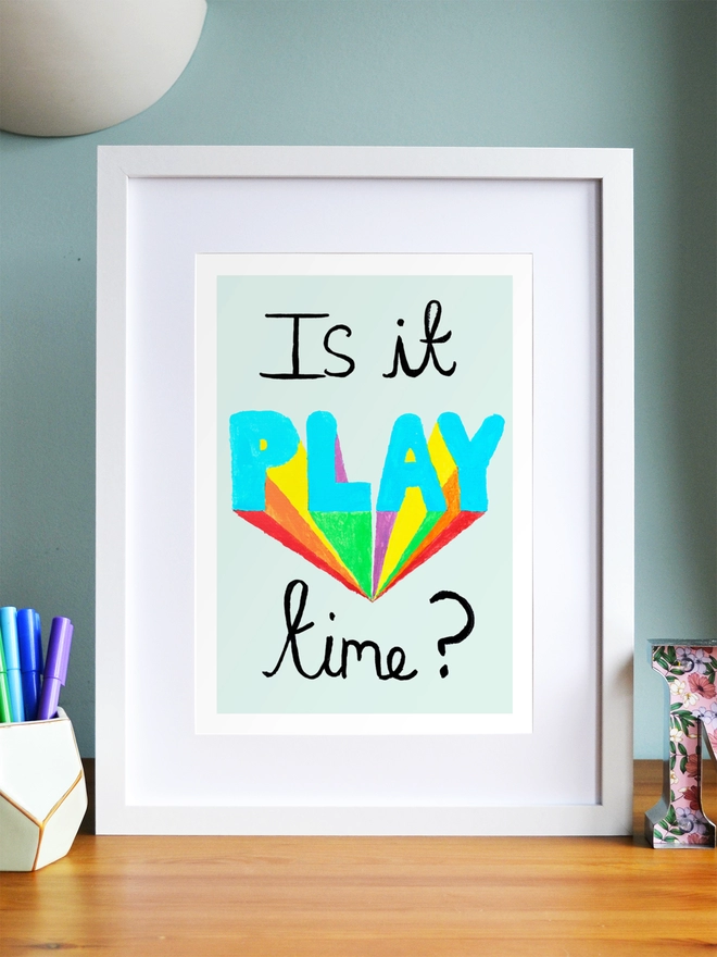 Art print saying 'Is it play time?' in a white frame on a child's desk