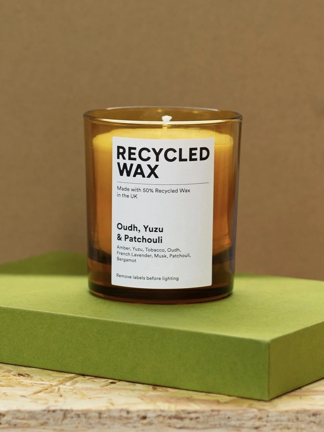 Glass Scented Candle. the oudh, yuzu and patchouli glass scented candle stands on a green box against a brown background, housed in an amber glass jar with a single wick. a white label reads "recycled wax, made with 50% recycled wax in the uk", followed by the scent name and note list of amber, yuzu, tobacco, oudh, french lavender, musk, patchouli and bergamot. the warm amber glass gives the candle a simple, understated finish.