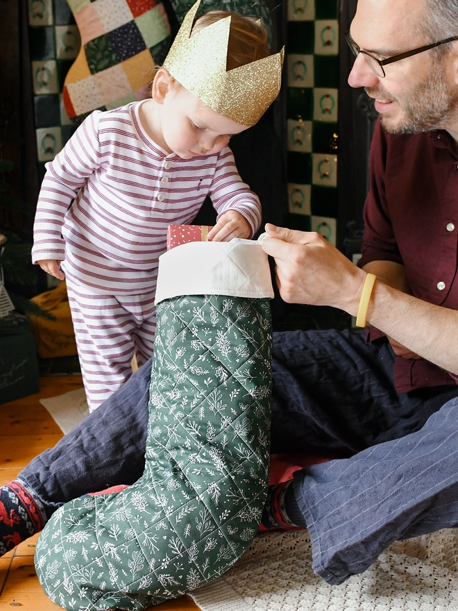 A young toddler wearing striped pyjamas and a golden crown looks into the top of a Christmas stocking being held by his Dad.