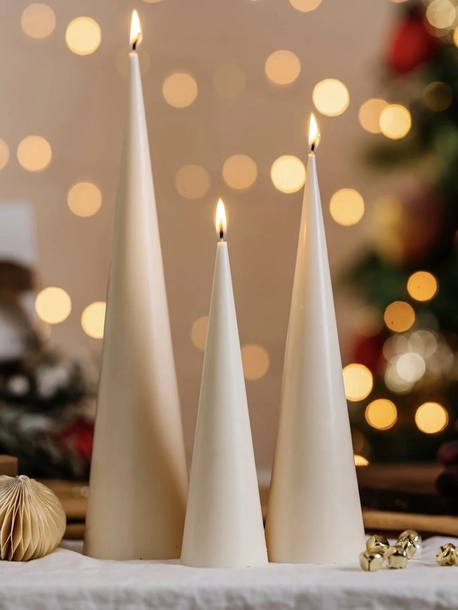 White Christmas Tree Cone Candles (Set Of 3). Three lit cream cone candles in graduating heights, styled on white linen with a paper bauble and gold bells against festive bokeh lights. The sleek minimalist shape makes an elegant Christmas centrepiece.