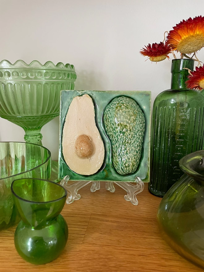 Decorative eye-catching avocado feature tile – three-dimensional, very realistic and incredibly tactile - displayed on a mantelpiece alongside antique green glass. 