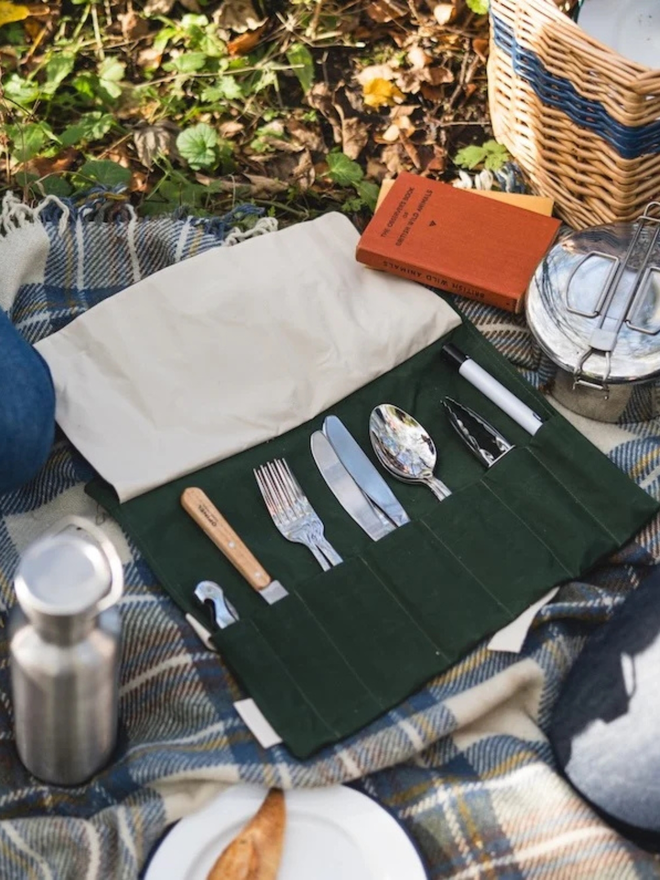 'mess kit' cotton cutlery and utensil roll. a pack of picnic utensils in a cotton roll, open and pictured on a chequered picnic rug.