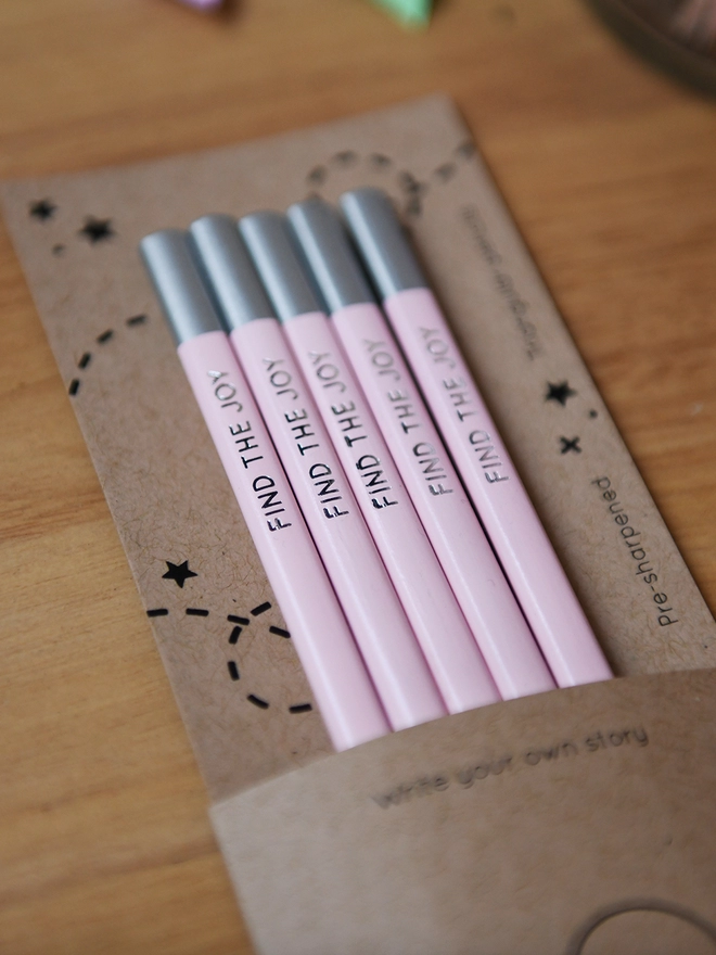 Five pink pencils with the words Find The Joy along the side of each one, are tucked into cardboard packaging on a wooden desk.
