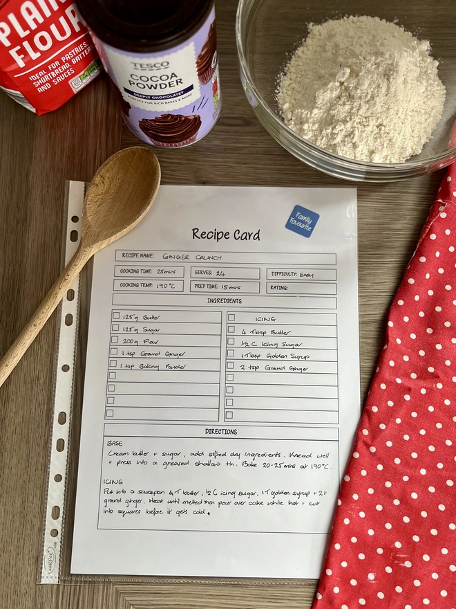 A4 recipe card page in a clear protective sleeve, lying on kitchen bench with wooden spoon, red polka-dot apron and baking ingredients.
