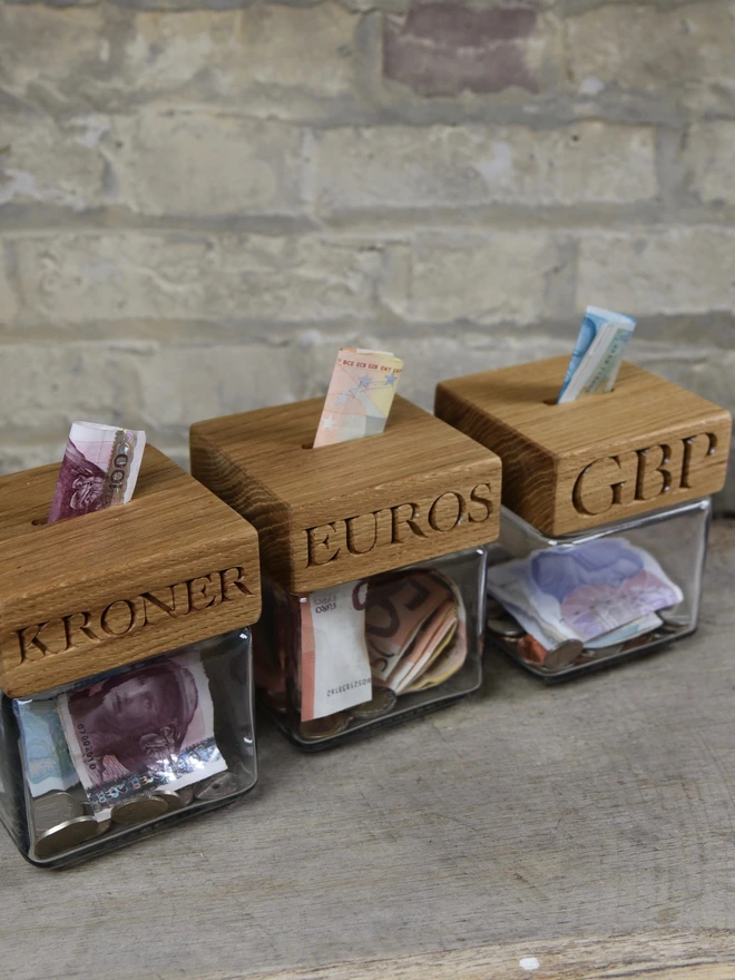 Three oak money jars engraved KRONER, EUROS and GBP, with bank notes pushed into the coin slots.