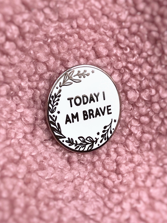A round white enamel pin with a floral design and the words "Today I Am Brave" is pinned to pink fabric.