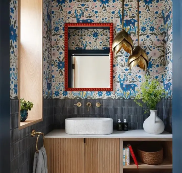 Over-sized red bobbin mirror with a square turned-bead frame mounted above a bathroom vanity, set against patterned blue floral wallpaper with brass pendant lights and a stone basin sink.