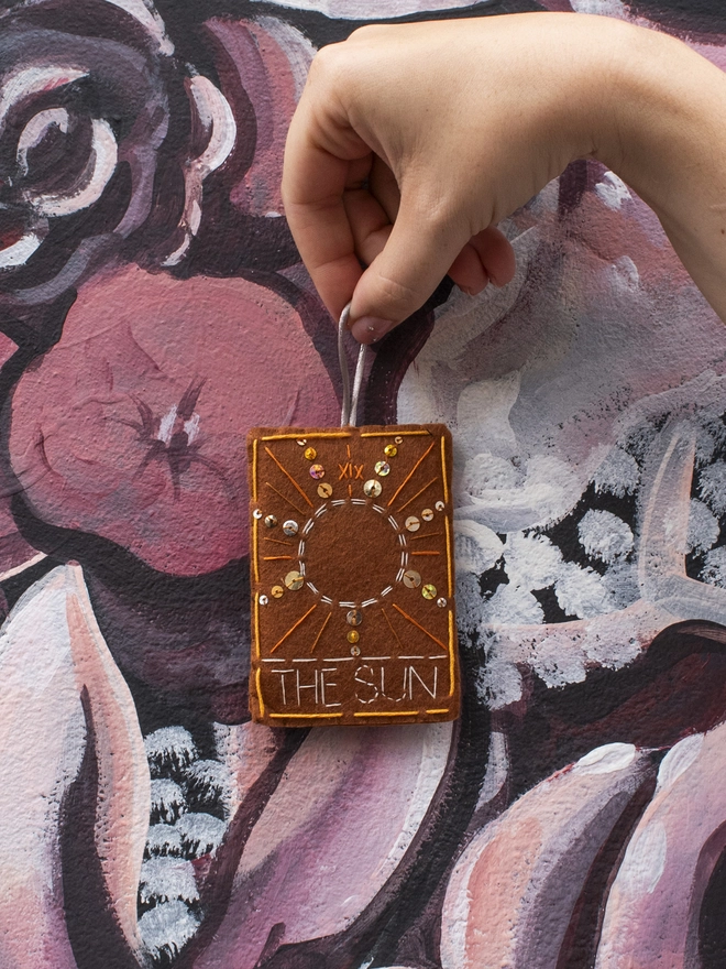 A handcrafted felt ornament with sequins and embroidery depicting "The Sun" tarot card.