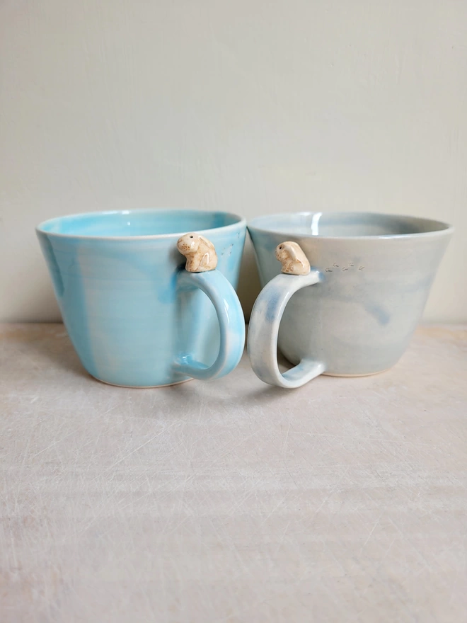 Two ceramic cups one grey and one blue with a little brown bunny rabbit on the handle 