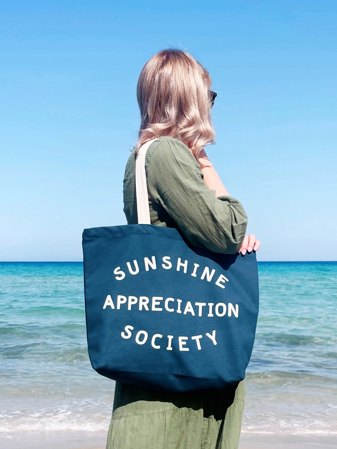 model carrying a large blue canvas tote bag with the words sunshine appreciation society on in front of the sea