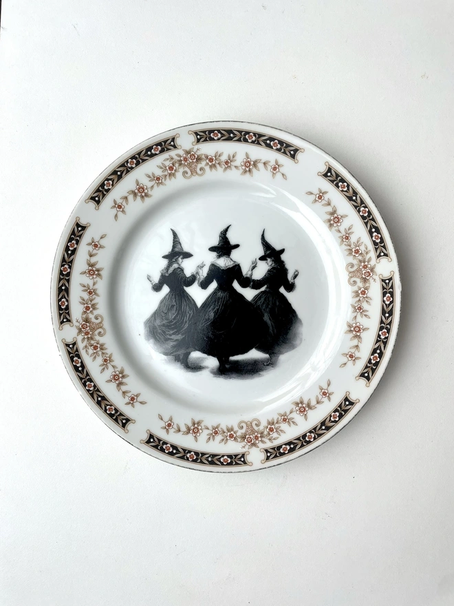 a coven of 3 witches dance hand in hand, the image is victorian in black and white and is printed directly onto an ornate vintage plate