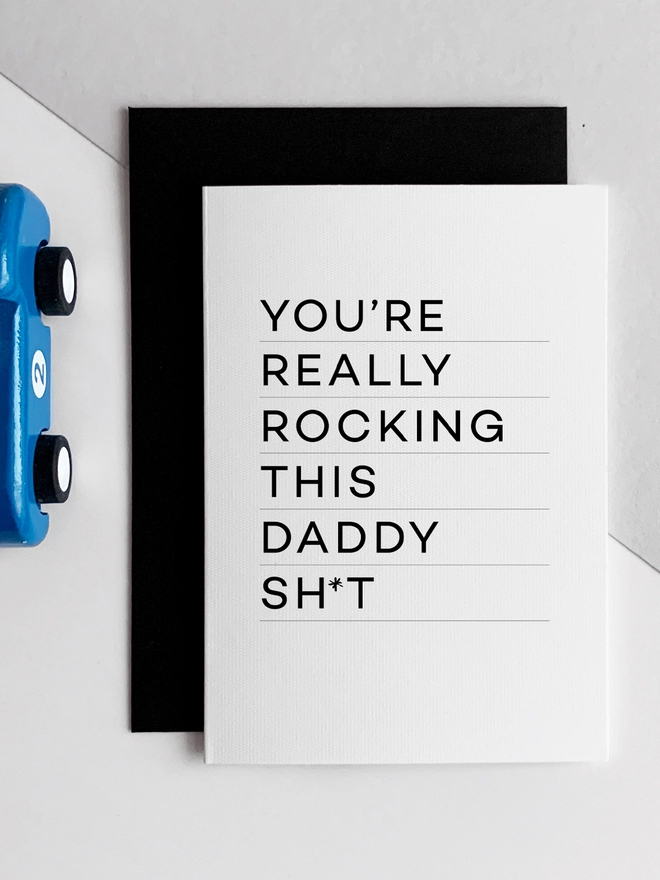 Rocking Daddy Birthday Card overlapping a black envelope next to a blue racing car.