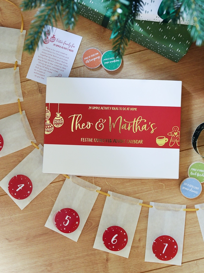 A white advent calendar box with a red sleeve lays on a wooden floor with white numbered envelopes beside it.