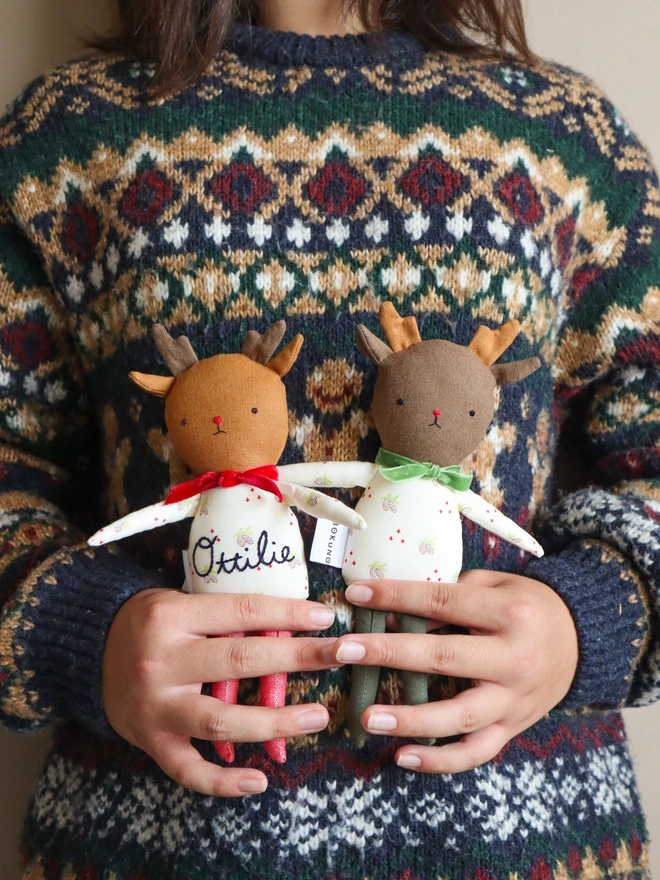 Two handmade reindeer dolls on a green background. Each doll has soft brown antlers, embroidered facial features, and wears a festive white outfit with a pinecone print. One doll has red legs and a red velvet bow; the other has green legs, a green bow, and the name “Ottilie” embroidered on its body. Both have a "BOKUNO" fabric tag.