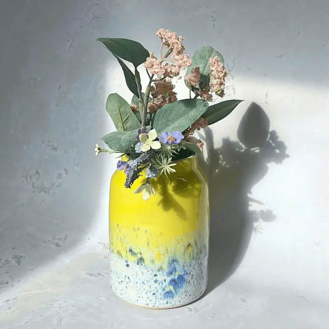 Shoreline Large Vase, a colourful vase placed on a stone surface in front of a plain backdrop. It is accompanied by flowers. 