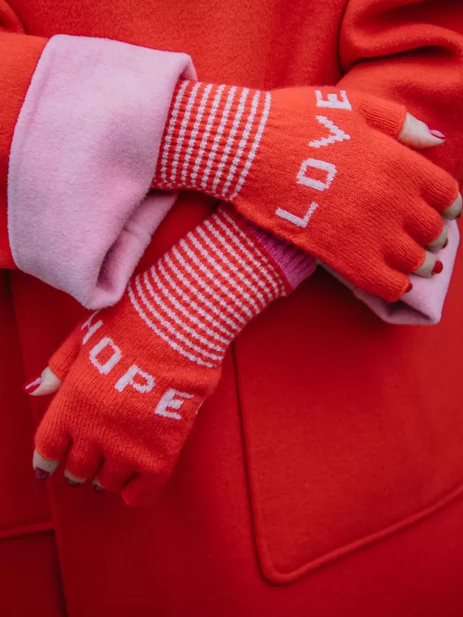 A classic red fingerless knitted glove with the words LOVE and HOPE in dusky pink and stripe cuff with a bubblegum pink edge detail. Made from 100% Lambswool with Wool spun in Yorkshire. 