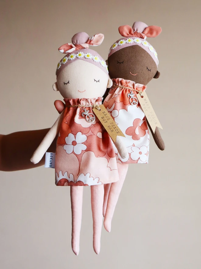 Two handmade fabric dolls called Peace Wishing Flower Child Dolls, each wearing peach and white retro floral dresses, daisy headbands, and bow topknots. They have serene embroidered faces, peace sign charms around their necks, and tags reading “Make Peace Not War.” One doll has a light complexion and the other has a darker complexion, both held side by side against a plain background.