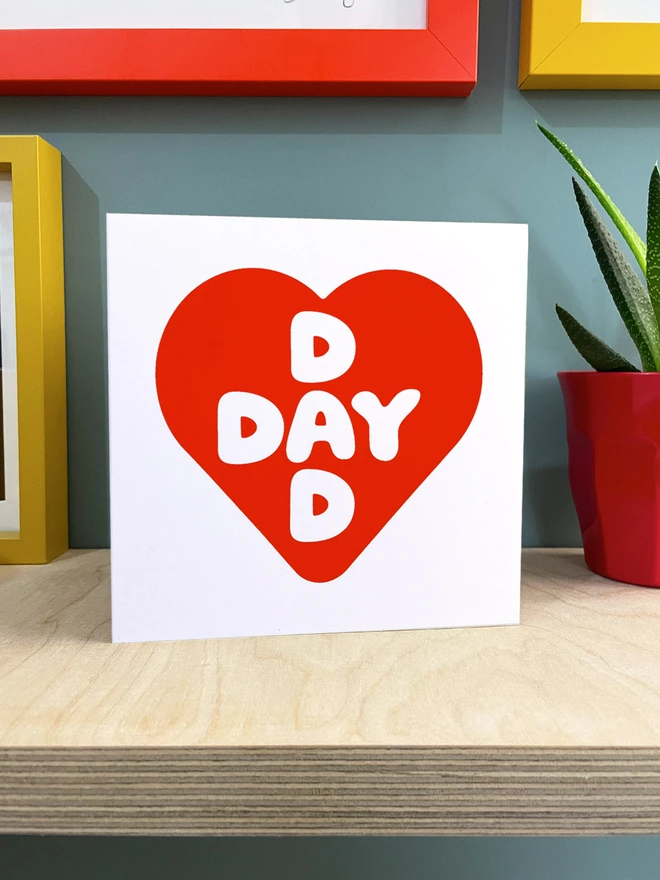 red screenprinted heart contains intersecting words Dad and Day, a square card stood on a ply shelf with hint of plant and other colourful frames.