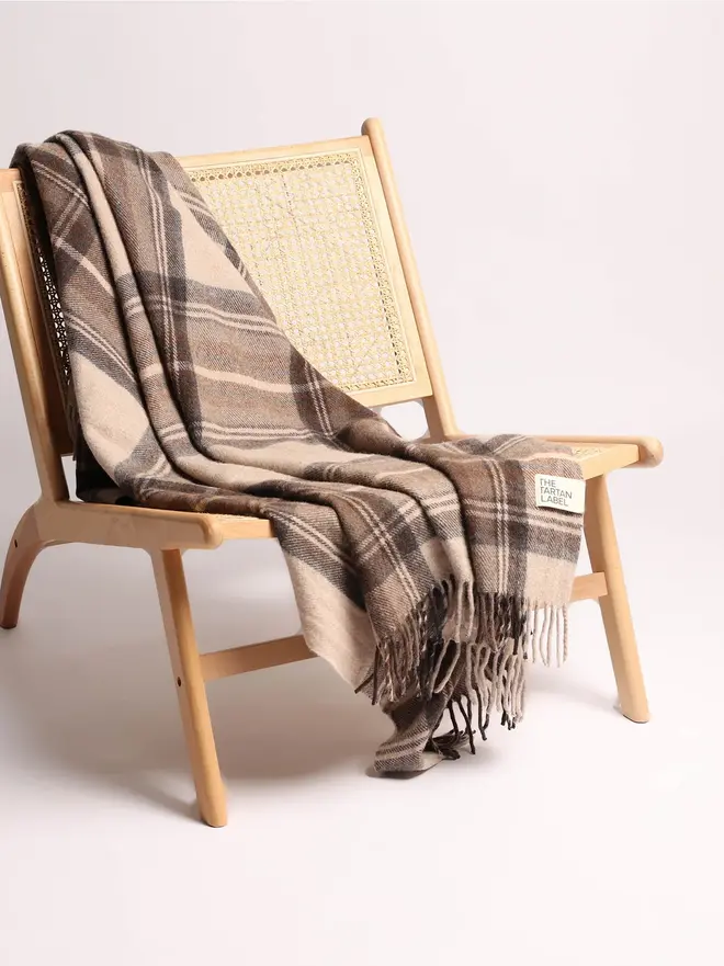 Stewart Natural Dress Blanket, a brown blanket draped over a wooden chair on a white surface against a plain backdrop. 