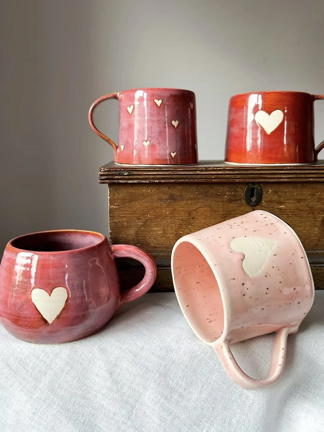Love Heart Mug Raspberry, a red heart mug on a wooden surface in the background accompanied by other mugs. 