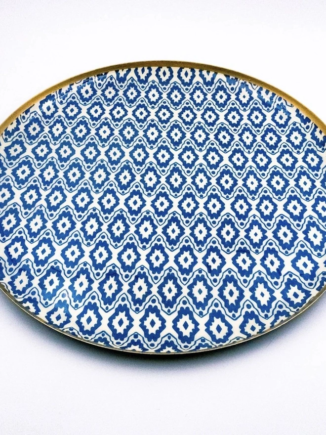 Large Enamel Circular Tray. A large round tray decorated with blue and cream geometric ikat-style print and finished with a gilded gold rim. The glossy serving or display tray adds bold pattern to a coffee table, ottoman or sideboard.