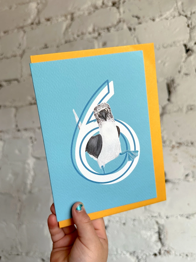 a bright blue card with a big white number 6 and an illustrated blue footed booby with a bright yellow envelope