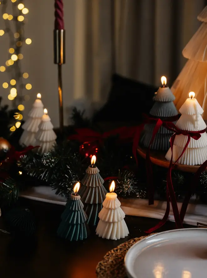 Forest Green Christmas Tree Warm Candles (Set Of 2). A Christmas table with lit tree candles in green, grey and cream, some tied with burgundy velvet bows, among pine garland and a brass taper holder. The mixed colourways make a warm, layered holiday display.
