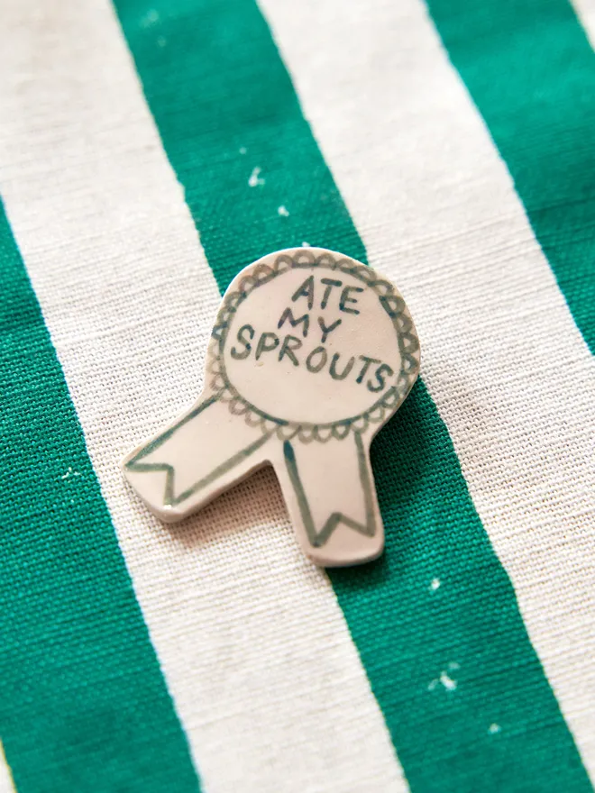 'Ate my Sprouts' hand-painted on a ceramic Christmas award badge in green, atop a striped green and white tablecloth