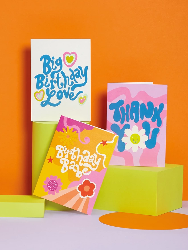 A trio of birthday and a thank you cards sit on lime green plinths against an orange background, the Raspberry Blossom cards have a vibrant 1970’s inspired design and a colour palette to match too 