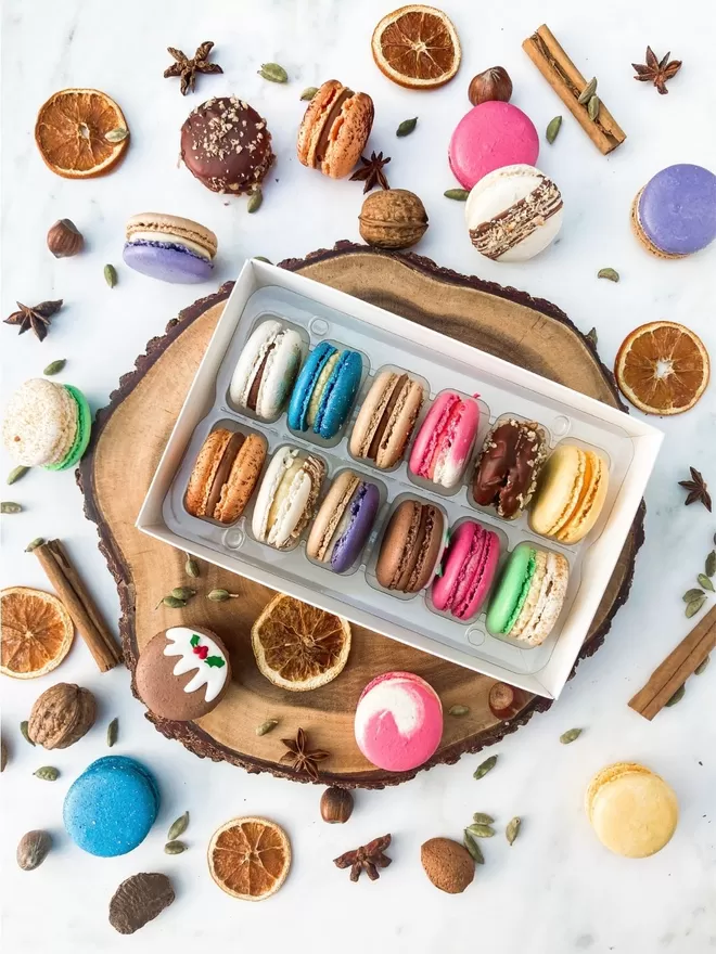 assorted colourful Christmas macarons in a white box on a wooden plate decorated with dried oranges, mixed spices and nuts