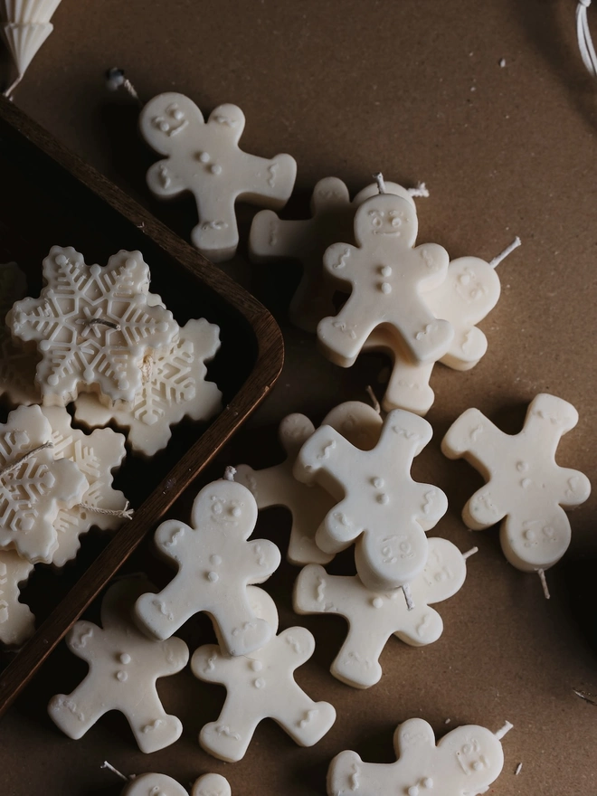 White Gingerbread Candles (Set Of 2). A pile of cream gingerbread man candles with embossed smiling faces, buttons and icing details, next to a tray of snowflake candles. The cute handmade candles add a sweet, nostalgic touch to Christmas décor.