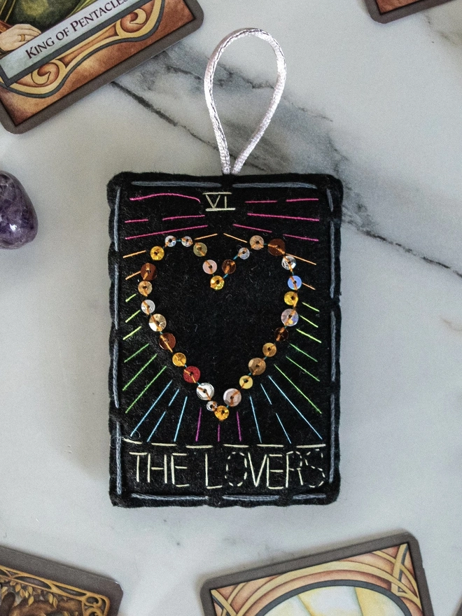 A handcrafted felt ornament with sequins and embroidery depicting "The Lovers" tarot card. Colourful embroidery with a heart shape. 