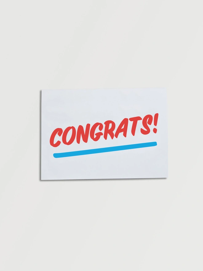 Folded A5 piece of paper that says CONGRATS! in red fun slanted typography on an angle with a blue underline