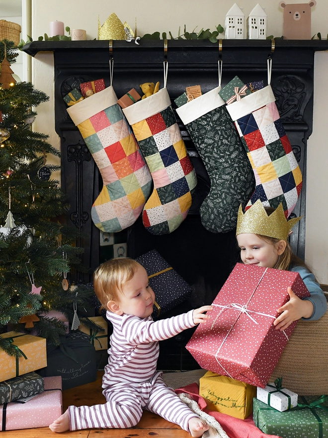 Two young children holding large wrapped gifts sit in front of a fireplace where four patchwork stockings hang. A Christmas tree stands beside them.
