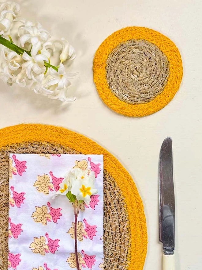 Close up of a seagrass coaster with yellow rings, shown on a light surface with small white flowers. 