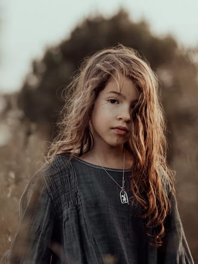 Girl stands outdoors in nature with warm sunlight, she looks at the camera wearing a silver chain with gold and silver pendant suspended on chain