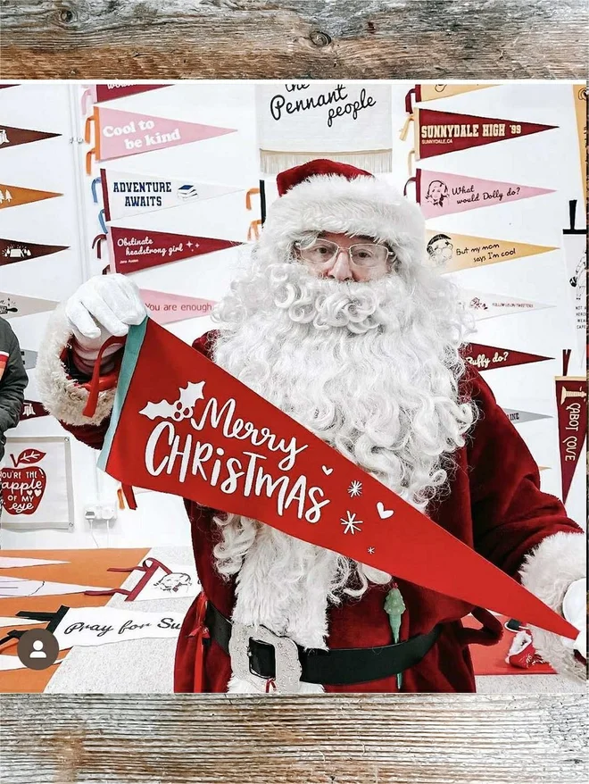 a red and white festive felt flag with the words Merry Christmas printed on it, it is being held by Father Christmas Santa Claus