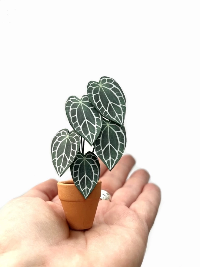A miniature replica Anthurium Crystallinum paper plant ornament in a terracotta pot sat on the palm of a hand