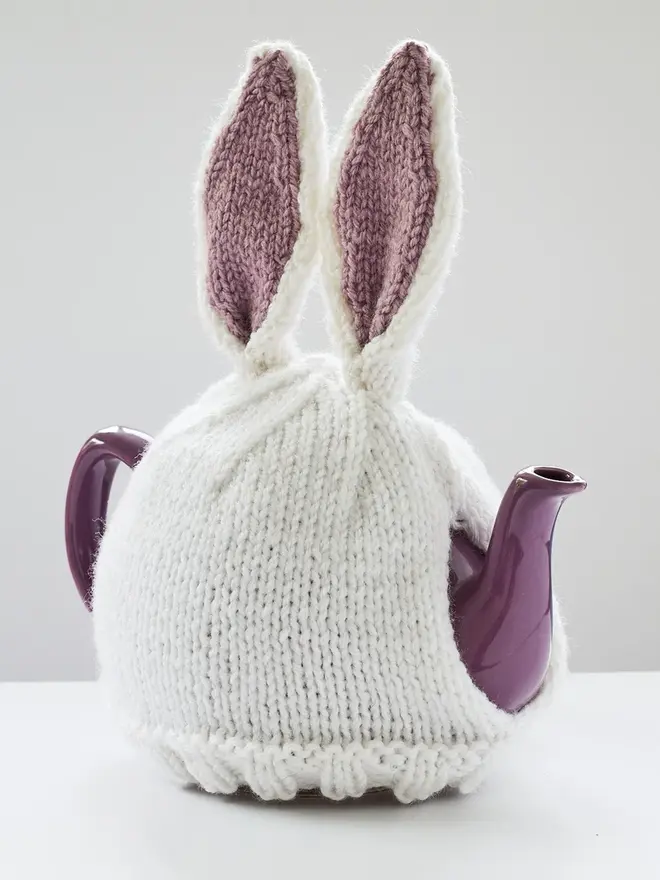 Hand Knitted Bunny Rabbit Tea Cosy, a rabbit bunny tea cosy sitting on a grey surface against a white backdrop. It fits over a purple tea pot. 