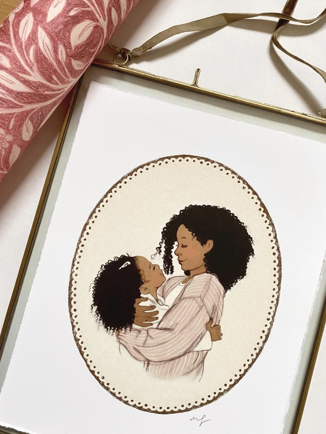 a phot of a print in a glass frame with a velvet ribbon and brass edging. illustration is on a black mother hugging her black daughter on a cream background with a hand drawn decorative sepia border