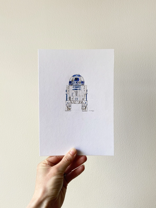 Hand drawn and painted watercolour illustration of R2D2 with a detailed yet organic and slightly loose style. The painting is a small illustration sitting on a white background. A white woman’s hand and arm can be seen holding up the A5 print between finger and thumb against an off white wall behind. 