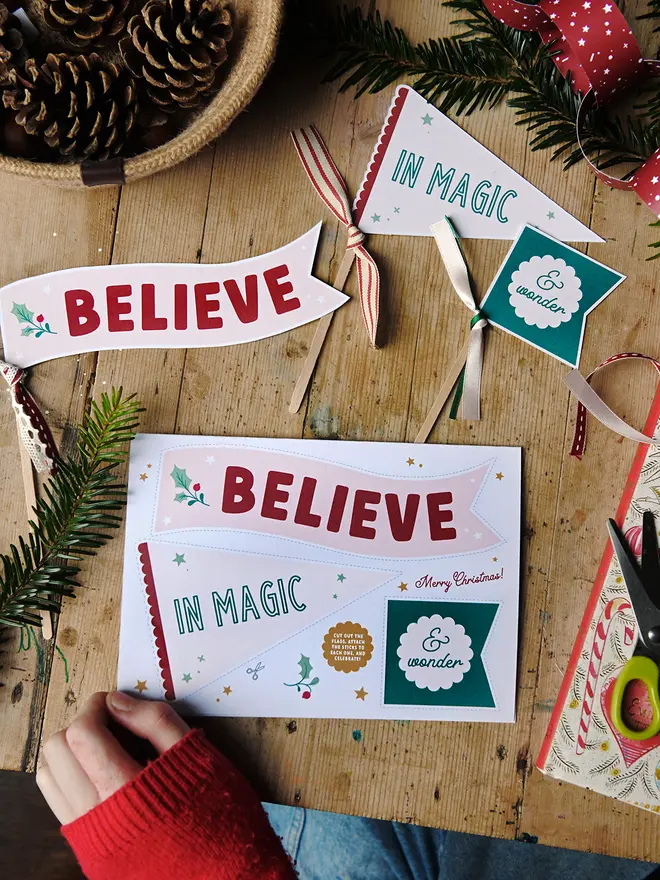 A Christmas card featuring three Christmas flags that read "Believe in magic & wonder" lays on a wooden table with various Christmas greenery, books & decorations around it.