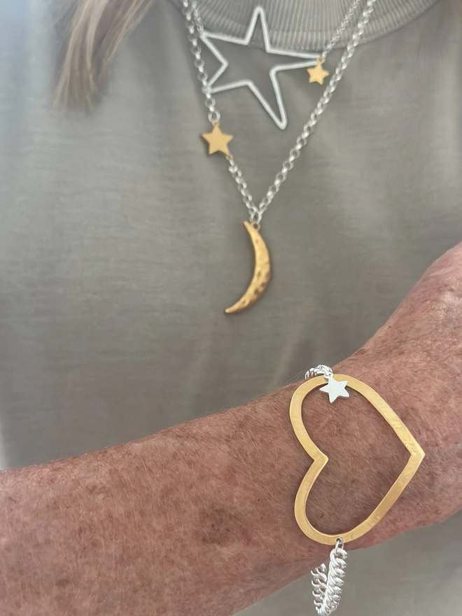model wears a large open heart charm in gold plate on a chunky curb chain bracelet with a small silver star charm. 