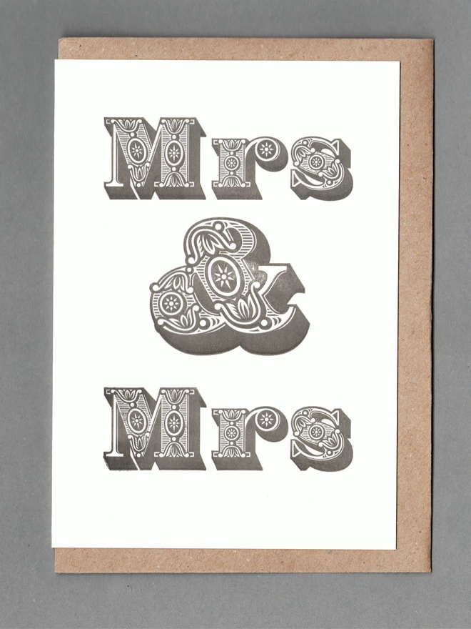 Black text reading 'Mrs & Mrs' on white card with brown envelope behind on grey background